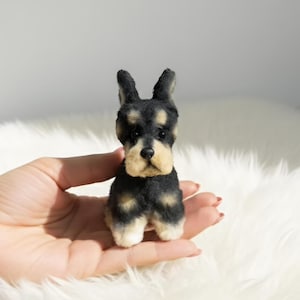 May include: A miniature felted dog figurine in black, tan, and cream. The small dog has upright ears and a detailed face. The figurine is held in a hand against a white, fluffy background. The dog is a miniature Schnauzer.