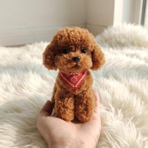 May include: A small, brown toy poodle with curly fur and a red bandana. The dog is sitting on a fluffy white surface. The dog is being held in a person's hand.