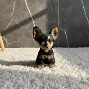 May include: A handcrafted miniature Chihuahua dog figurine. The dog is black and tan with large, upright ears. It is sitting on a white, fluffy surface, with a dark gray background. The figurine is made of felted wool.