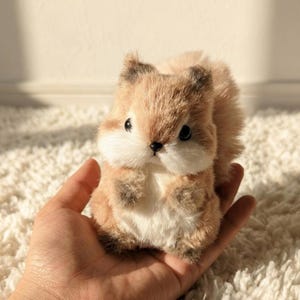 May include: A small, plush squirrel figurine with soft, light brown and white fur. The squirrel has large, dark, round eyes, a tiny black nose, and a bushy tail. It is designed with a rounded body and short limbs, featuring a dense, textured appearance.