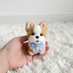 May include: A small, plush dog figurine with a white and tan coat, pink-lined ears, and a blue gingham bow tie. The toy dog has a black nose and eyes, and is approximately 7.6 cm tall. The figurine is designed to look like a puppy.