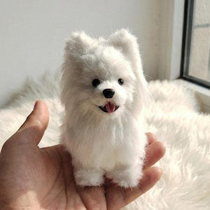 May include: A small, fluffy white dog figurine with black eyes and a pink tongue. The toy dog has long, soft fur and is held in a hand. The dog is a miniature replica of a real dog breed.