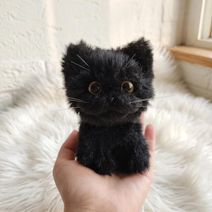 Op de afbeelding: Een kleine, zwarte pluche kat met grote, ronde, amberkleurige ogen en lange snorharen. De kat wordt in een hand gehouden, wat de zachte, getextureerde vacht laat zien. De speelgoedkat heeft een schattig, expressief gezicht.