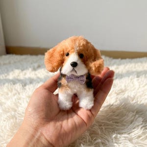 Puede incluir: Un juguete de peluche con forma de perro beagle con pelaje marrón, blanco y negro. El juguete tiene orejas caídas y una pajarita a cuadros morada. El perro es pequeño.