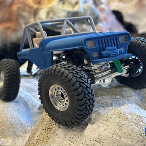 Könnte beinhalten: Ein blaues Spielzeug-Geländefahrzeug mit großen, schwarzen Reifen und einem Überrollkäfig. Der Miniatur-LKW hat ein detailliertes Fahrgestell und eine Federung, mit einer grünen Komponente. Das Fahrzeug steht auf einer felsigen Oberfläche.