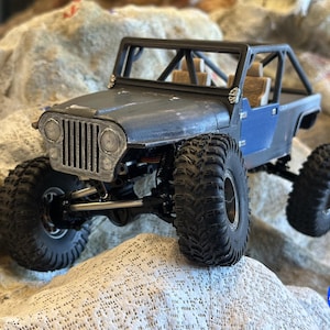 Könnte beinhalten: Detailliertes Modell eines blau-grauen Geländewagens mit großen, strukturierten Reifen. Das Miniatur-Jeep-ähnliche Fahrzeug hat einen Überrollkäfig und befindet sich auf einer felsigen Oberfläche. Das Bild enthält ein Logo in der unteren rechten Ecke.