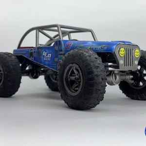 May include: A blue and silver toy off-road vehicle with large, black tires. The vehicle has a roll cage and the text "4.0 Micro" on the side. The front features a silver grill with smiley face headlights.
