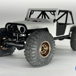 Könnte beinhalten: Ein weiß-schwarzes Miniatur-Geländefahrzeug mit großen, strukturierten Reifen und goldfarbenen Felgen. Das Modell hat einen Überrollkäfig und ein detailliertes Fahrgestell, das ein robustes Design zeigt. Das Fahrzeug ist ein Modell.