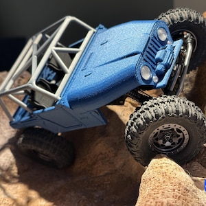 以下が含まれることがあります： 青とシルバーのオフロード玩具車。大きなブロックタイヤを装着。 ロールケージがあり、岩の表面に配置され、そのクライミング能力を示しています。 画像には「MICRO 4.0」の文字が含まれています。