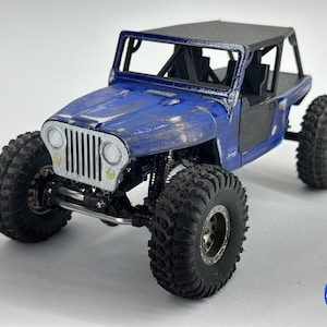 以下が含まれることがあります： 青と黒の玩具ジープモデル。 大きなテクスチャードタイヤが付いています。 モデルにはロールケージ、黒い屋根、詳細なフロントグリルがあります。 側面には「Jeep」の文字が見えます。 モデルは白い表面にあります。