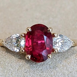 May include: A gold ring featuring a large oval-cut ruby center stone flanked by two pear-shaped diamonds. The ruby is a deep red color, and the diamonds are clear. The ring is set on a gold band. Text on the image says "ShineToDivine".
