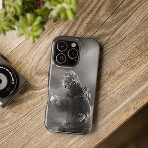 May include: A smartphone case featuring a black and white graphic of the monster Godzilla. The design shows the creature in a textured, dark grey scale with sharp dorsal fins, standing against a cloudy, monochromatic background on the phone back.