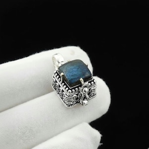 May include: A silver pendant with a square, blue gemstone. The pendant has an ornate, box-like design with intricate patterns and a small loop for a chain. The gemstone has a dark blue color.