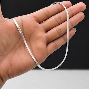 Op de afbeelding: Een zilverkleurige visgraatketting met een plat, flexibel en glad schakelontwerp. De metalen ketting heeft een reflecterend oppervlak en een consistente breedte over de gehele lengte, waardoor een eenvoudige lus wordt gevormd.
