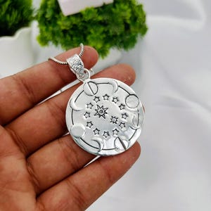 May include: A round silver-toned pendant featuring an embossed design of moon phases encircling a central arrangement of stars. The pendant is attached to a silver-toned chain with a textured bail, showcasing a polished metallic finish throughout.