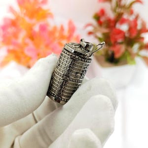 Puede incluir: Un colgante cilíndrico de metal en tono plateado con un patrón de rejilla texturizado y detalles decorativos de volutas. El colgante tiene una pequeña anilla en la parte superior para su sujeción y un acabado pulido con intrincados detalles.