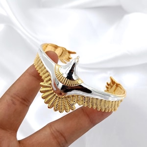 May include: A two-tone cuff bracelet with a gold and silver eagle design. The bracelet features intricate detailing, with a textured gold base and a silver eagle head. The bracelet is held by a hand against a white background.