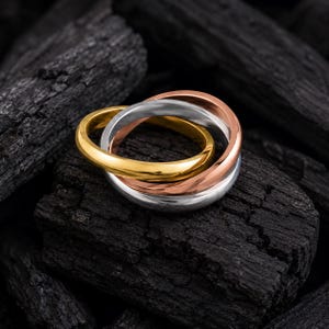 May include: A tri-color rolling ring featuring three interlocking bands in polished yellow gold, rose gold, and silver-toned metal. The smooth, rounded bands are connected in a continuous loop design, showcasing a reflective metallic finish.