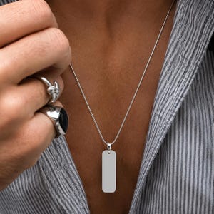 Op de afbeelding: Een zilverkleurige ketting met een gladde, rechthoekige verticale hanger aan een dunne ketting. De hanger heeft afgeronde hoeken en een gepolijst, reflecterend oppervlak. De ketting wordt gedragen over een grijs-wit gestreept overhemd.