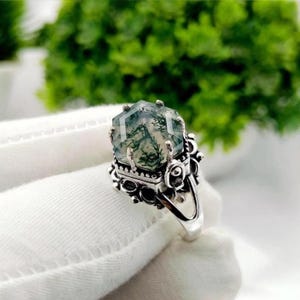 Puede incluir: Un anillo de plata con una piedra preciosa de ágata musgosa hexagonal con inclusiones dendríticas de color verde oscuro. El engaste presenta un intrincado trabajo en metal, incluyendo pequeñas esferas decorativas y recortes.