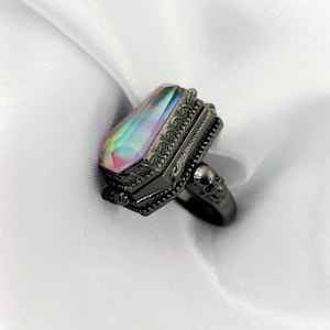 May include: A black ring featuring a coffin-shaped, iridescent gemstone. The ring has detailed embellishments around the gem and a skull motif on the band. The gemstone displays a range of colours, including pink, green, and blue. The ring is set against a white backdrop.