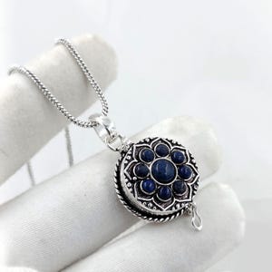 Puede incluir: Un collar con colgante plateado con un diseño circular inspirado en flores. El colgante presenta una piedra azul oscuro central rodeada de piedras azules más pequeñas. La cadena del collar es de plata fina y texturizada.