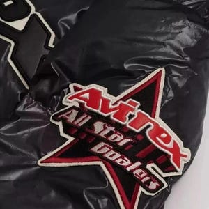 Puede incluir: Primer plano de una chaqueta negra con acabado brillante. Presenta un parche bordado rojo y blanco con el texto "Avirex All Star Dealers" en forma de estrella. La chaqueta tiene una textura acolchada.