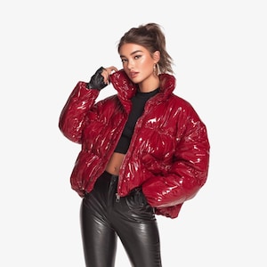 Puede incluir: Chaqueta acolchada roja brillante con cuello alto y cierre de cremallera. La chaqueta tiene un acabado brillante y un diseño abultado. La modelo lleva guantes negros y pantalones negros. El fondo es blanco.