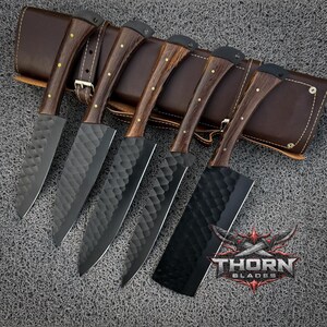 May include: A set of six kitchen knives with dark wooden handles and textured black blades. The knives are displayed on a brown leather sheath with a buckle. The logo "THORN BLADES" is visible.