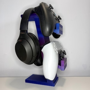 May include: A blue, purple, and black gaming headset and controller stand. The stand holds a black headset, a black controller, and a white controller. The stand is made of plastic and has a rectangular base.