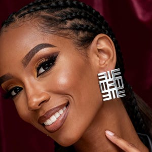 Puede incluir: Pendientes cuadrados plateados con diseño geométrico. Los pendientes los lleva una persona de piel oscura, pelo oscuro y top negro. El fondo es de color rojo oscuro.