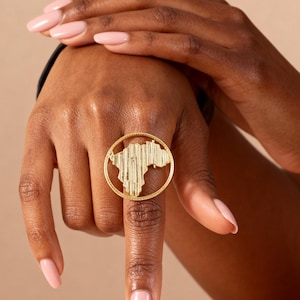 Può includere: Un anello dorato con un design dettagliato a mappa all'interno di una cornice circolare. L'anello è indossato su un dito, mostrando la sua intricata lavorazione. Il design dell'anello è il punto focale, evidenziando la sua forma e il suo stile unici.