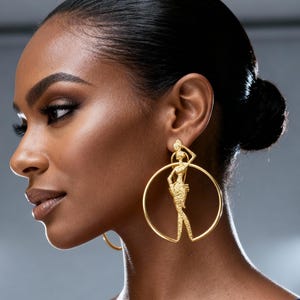 Può includere: Orecchini a cerchio dorati con un design a figura stilizzata. Gli orecchini presentano un cerchio circolare con una silhouette dettagliata di una persona. La figura è in posa con un braccio alzato. Lo sfondo è grigio tenue.