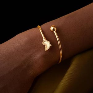 Puede incluir: Un brazalete abierto de color dorado. Un extremo presenta un mapa estilizado y texturizado de África, mientras que el otro extremo tiene una pequeña bola redonda. El brazalete se lleva en la muñeca.