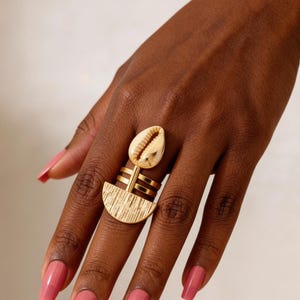 Op de afbeelding: Een goudkleurige ring met een centraal kauri-schelp element boven een getextureerde halfronde basis. Het ontwerp van de ring bevat drie parallelle horizontale banden die de schelp verbinden met het halfronde onderdeel op de vinger.