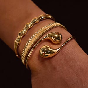 Puede incluir: Se muestran tres pulseras doradas en una muñeca. Las pulseras tienen diferentes diseños: una con una forma texturizada y orgánica, otra con un patrón acanalado y una tercera con extremos en forma de lágrima. Las pulseras son brillantes y reflectantes.