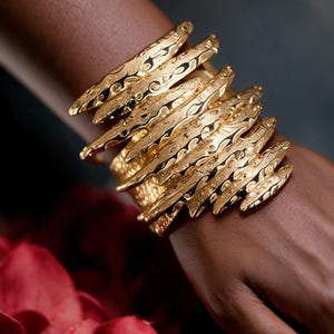 Puede incluir: Una pulsera brazalete ancha de color dorado con un diseño apilado de varias capas y una superficie texturizada y martillada. El metal tiene un acabado brillante y reflectante con hendiduras orgánicas irregulares en cada capa.