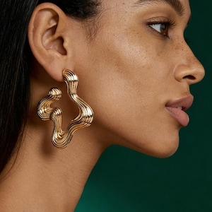 May include: Gold-coloured, abstract wave-shaped earrings. The earrings feature a textured, ridged surface and a unique, flowing design. The jewellery is shown against a dark green background, highlighting the metallic sheen of the earrings.