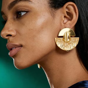 Peut inclure: Boucles d'oreilles dorées. Les boucles d'oreilles présentent un design en demi-cercle avec une section inférieure texturée en forme d'éventail et une section supérieure lisse plus petite. Un petit coquillage est centré sur le design. Les boucles d'oreilles sont portées par une personne aux cheveux et à la peau foncés.