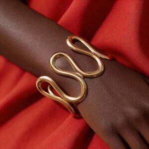 Puede incluir: Un brazalete dorado con un diseño abierto y fluido. El brazalete se envuelve alrededor de la muñeca, con una serie de bucles interconectados. La joya se muestra sobre un fondo rojo.