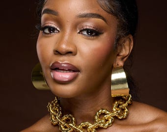 May include: A pair of wide, polished gold-tone hoop earrings with a smooth, curved surface, paired with a chunky, textured gold-tone chain link necklace featuring interconnected, twisted metal loops that sit close to the base of the neck.