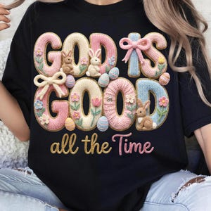 以下が含まれることがあります： 「GOD IS GOOD all the Time」の文字が3Dニットデザインで施された黒いTシャツ。パステルカラーのウサギ、卵、花、リボンがデザインされています。