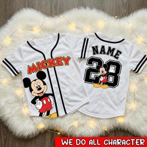 May include: Two white baseball-style shirts with black trim and buttons. One shirt features "Mickey" in red and yellow, with a Mickey Mouse graphic. The other has "NAME" above the number "28" and a Mickey Mouse graphic. "WE DO ALL CHARACTER" is at the bottom.
