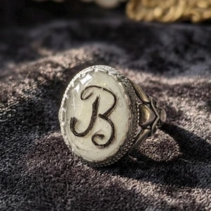 Può includere: Un anello d'argento con castone ovale in resina trasparente. All'interno del castone c'è uno sfondo bianco con la lettera nera "B". L'anello ha una fascia ornata e dettagliata.