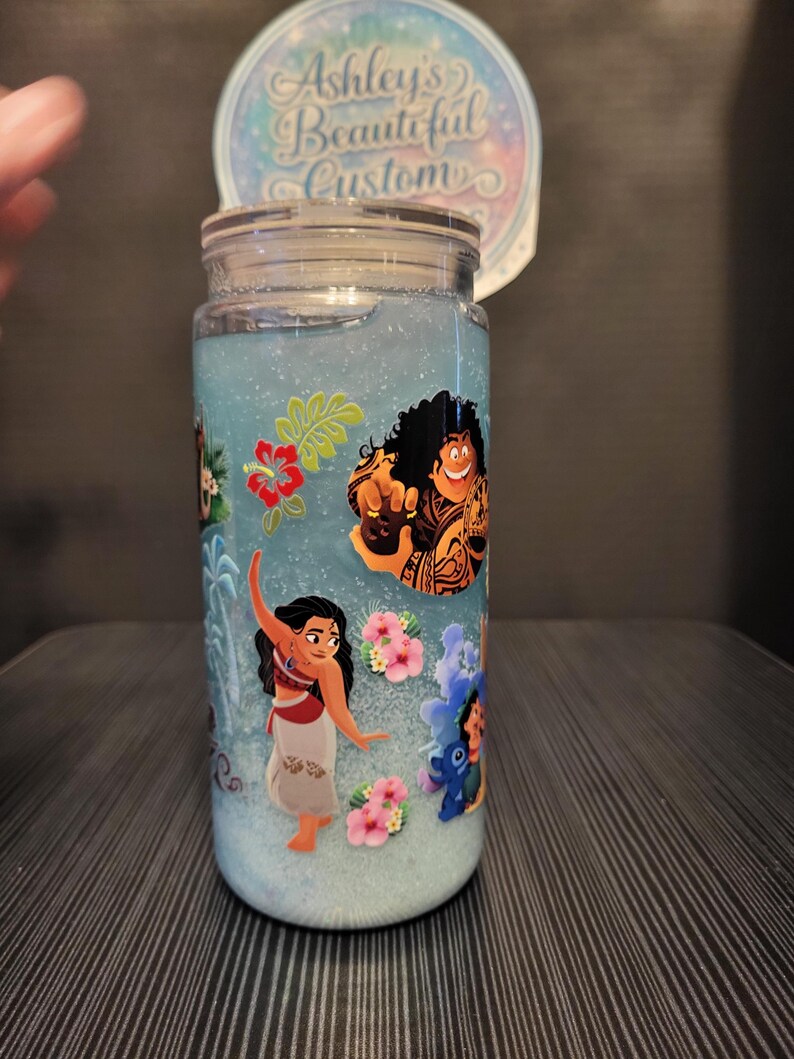 Puede incluir: Un tarro de cristal transparente con tapa, lleno de l&iacute;quido azul y purpurina. El tarro est&aacute; decorado con personajes de dibujos animados y flores rosas. Una pegatina circular en la parte superior dice "Ashley's Beautiful Custom".