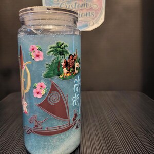Puede incluir: Un tarro de cristal transparente con tapa plateada, lleno de l&iacute;quido azul y elementos decorativos. El tarro presenta un dise&ntilde;o con personajes, un barco y flores rosas. El texto "Ashley's Beautiful Custom Creations" es visible en una pegatina en el fondo.
