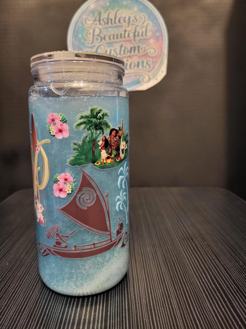 Puede incluir: Un tarro de cristal transparente con tapa plateada, lleno de l&iacute;quido azul y elementos decorativos. El tarro presenta un dise&ntilde;o con personajes, un barco y flores rosas. El texto "Ashley's Beautiful Custom Creations" es visible en una pegatina en el fondo.