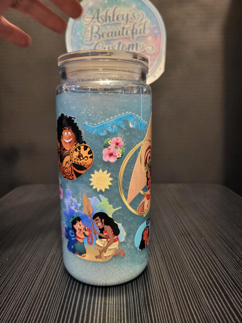 Puede incluir: Un tarro de cristal transparente con tapa, lleno de l&iacute;quido azul y purpurina. El tarro est&aacute; decorado con pegatinas de personajes de dibujos animados de colores. El texto "Ashley's Beautiful Customs" es visible en una pegatina circular.