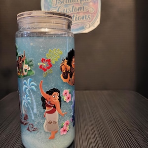 Puede incluir: Un tarro transparente con tapa, lleno de l&iacute;quido azul y purpurina. El tarro est&aacute; decorado con im&aacute;genes de personajes de la pel&iacute;cula de animaci&oacute;n "Vaiana" y flores tropicales. El texto "Ashley's Beautiful Custom Creations" est&aacute; en una pegatina en el fondo.