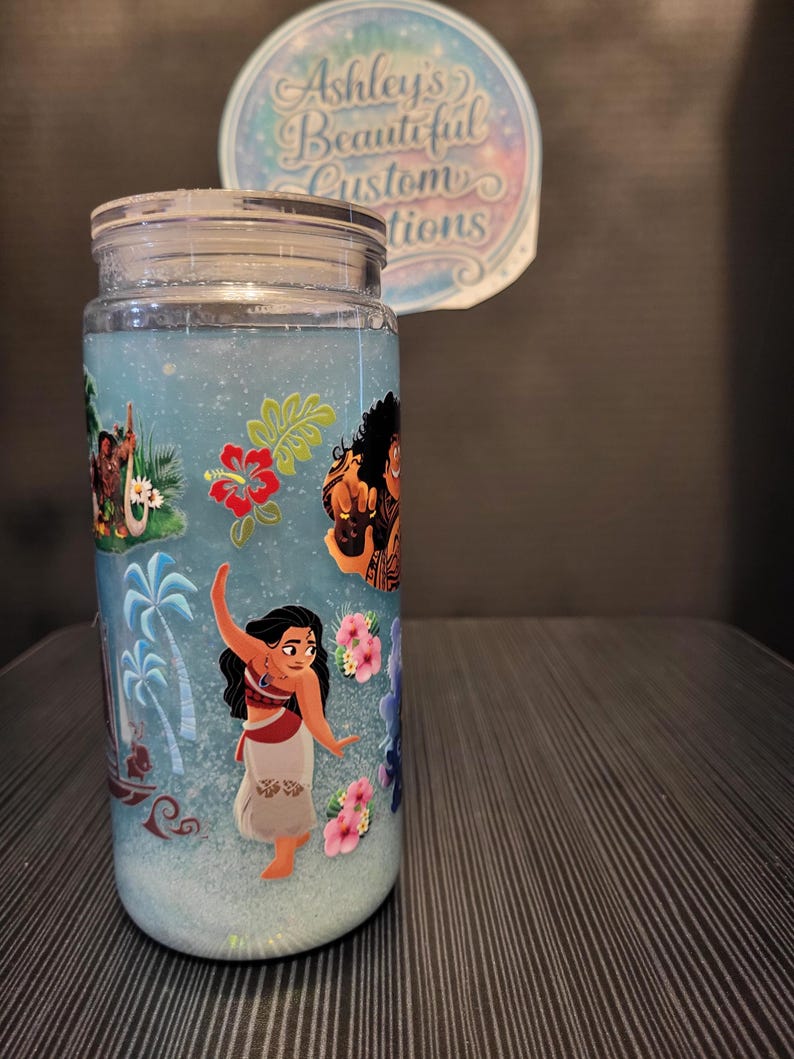Puede incluir: Un tarro transparente con tapa, lleno de l&iacute;quido azul y purpurina. El tarro est&aacute; decorado con im&aacute;genes de personajes de la pel&iacute;cula de animaci&oacute;n "Vaiana" y flores tropicales. El texto "Ashley's Beautiful Custom Creations" est&aacute; en una pegatina en el fondo.